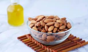 Close-up of almonds in a glass bowl next to a bottle of oil on a bamboo mat.