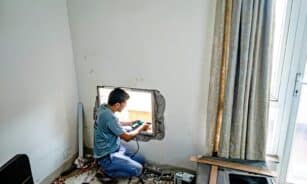 A man uses a drill to fit a window on an indoor construction site, focused on renovation work.
