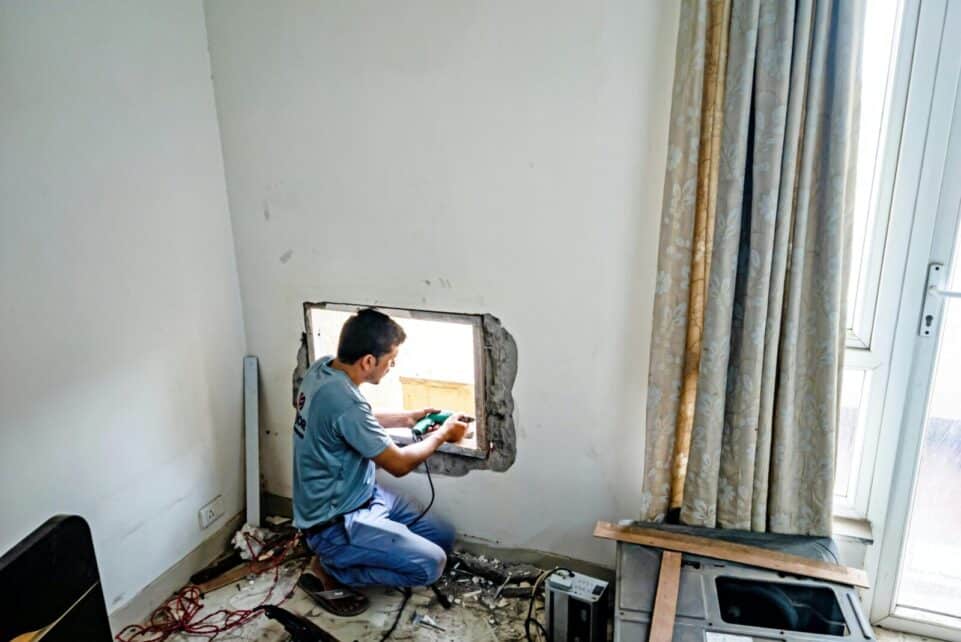A man uses a drill to fit a window on an indoor construction site, focused on renovation work.