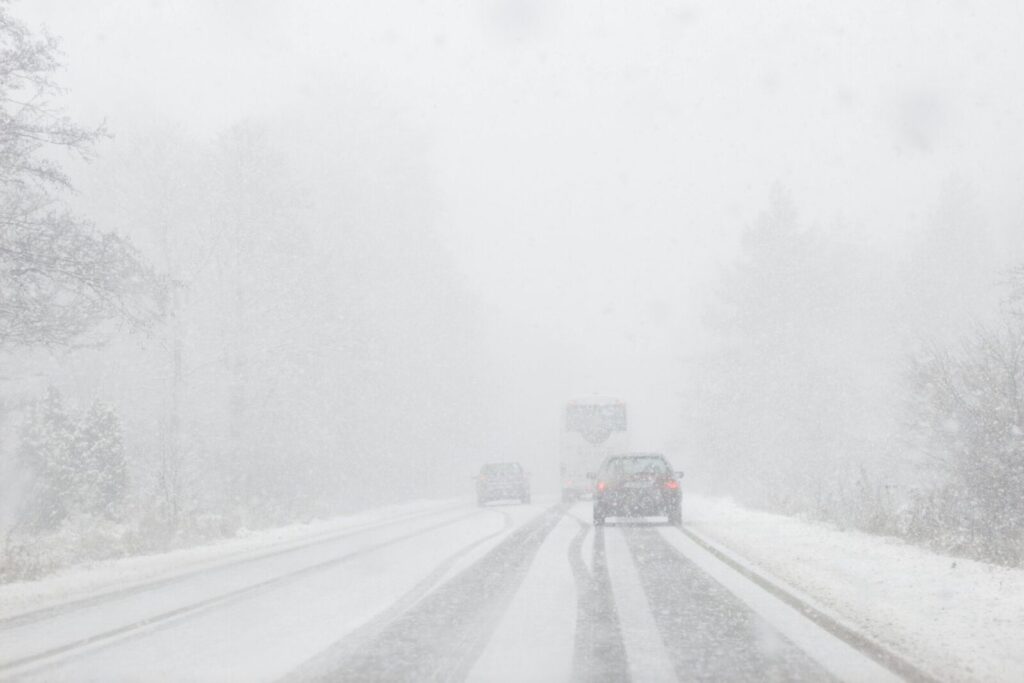 Heavy snowfall covers a road surrounded by a winter forest, with cars navigating the snowy conditions.