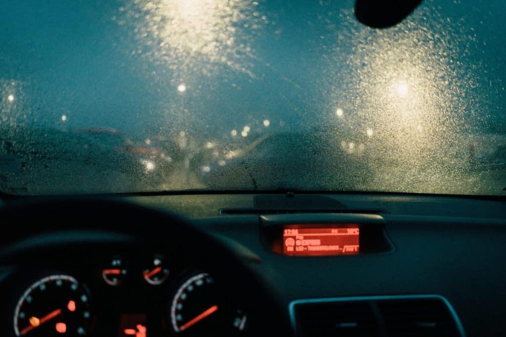 View from car interior with raindrops on windshield and blurred lights at night.