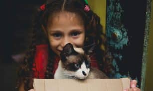 A joyful young girl with braids and a smiling face holding a box with a cat inside.
