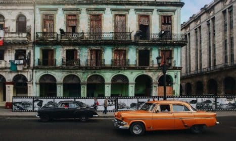 Classic cars passing by a decaying historic building in Havana, Cuba, showcasing vintage urban charm.