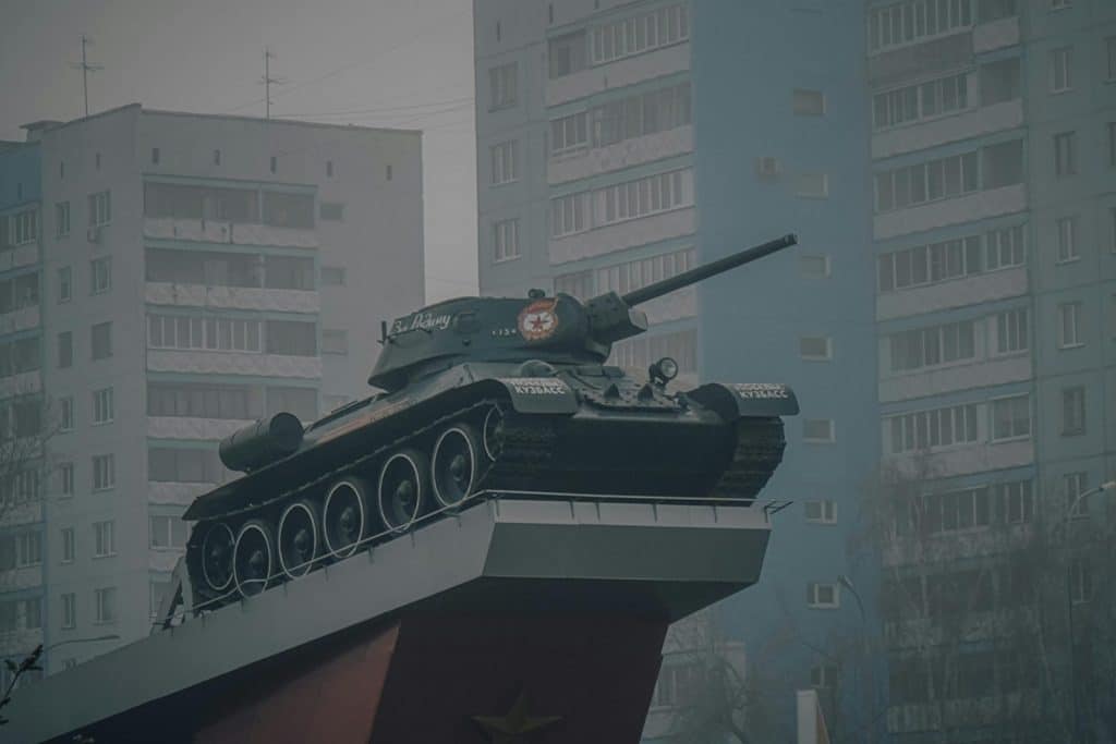 A military tank monument on a platform with residential buildings in the background.