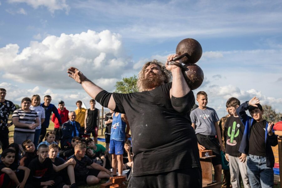 A strongman lifts a heavy dumbbell in an outdoor strength competition surrounded by a crowd.
