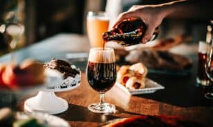 Sunlit scene with hand pouring dark beer into a glass. Includes baked goods and desserts on table.