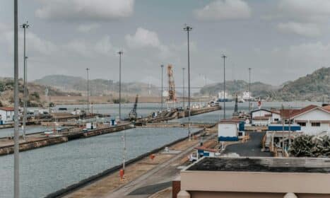 A panoramic daytime view of the Panama Canal locks with surrounding landscape and maritime infrastructure.