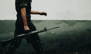 A soldier in camouflage walking with a rifle against a textured wall, day.