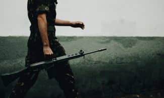 A soldier in camouflage walking with a rifle against a textured wall, day.
