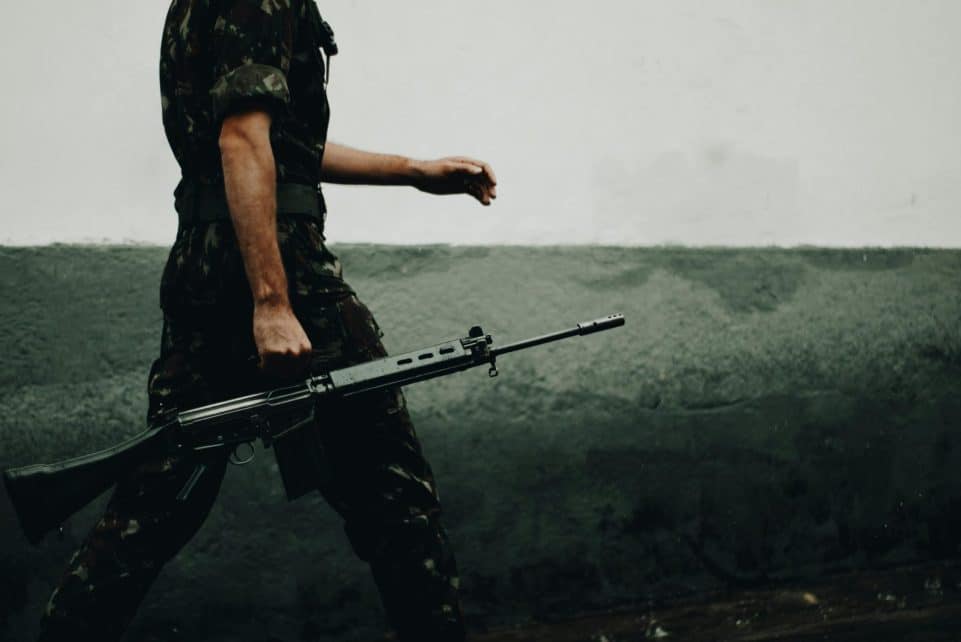 A soldier in camouflage walking with a rifle against a textured wall, day.