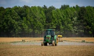 Tractor spraying fields with pesticides on a countryside farm in summer with a forest backdrop.