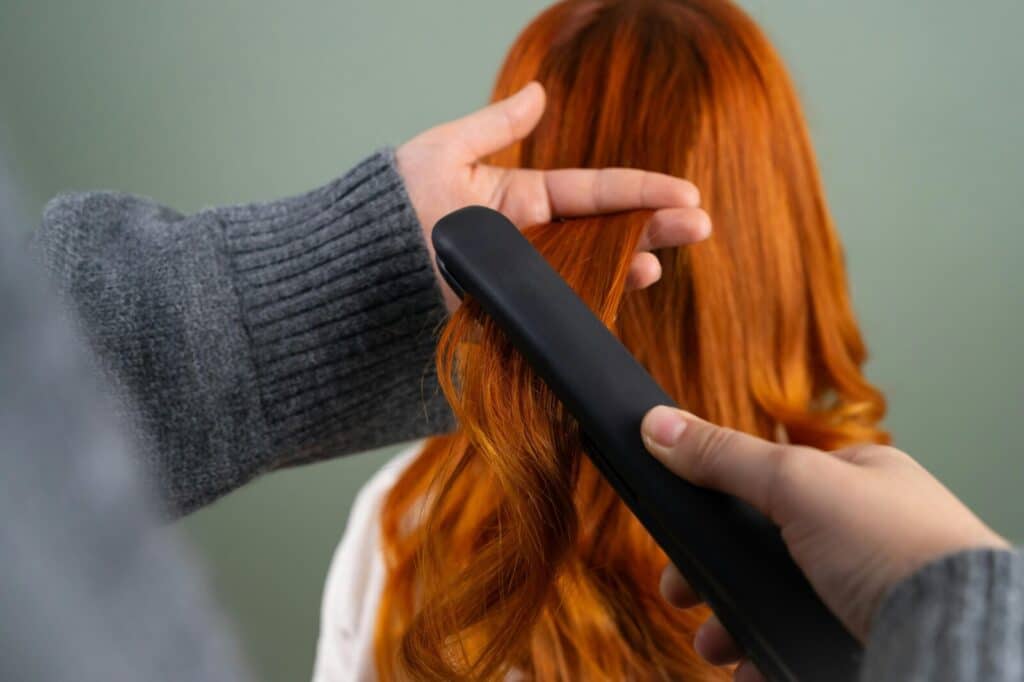 Hands styling long red hair using a flat iron indoors. Haircare and beauty concept.