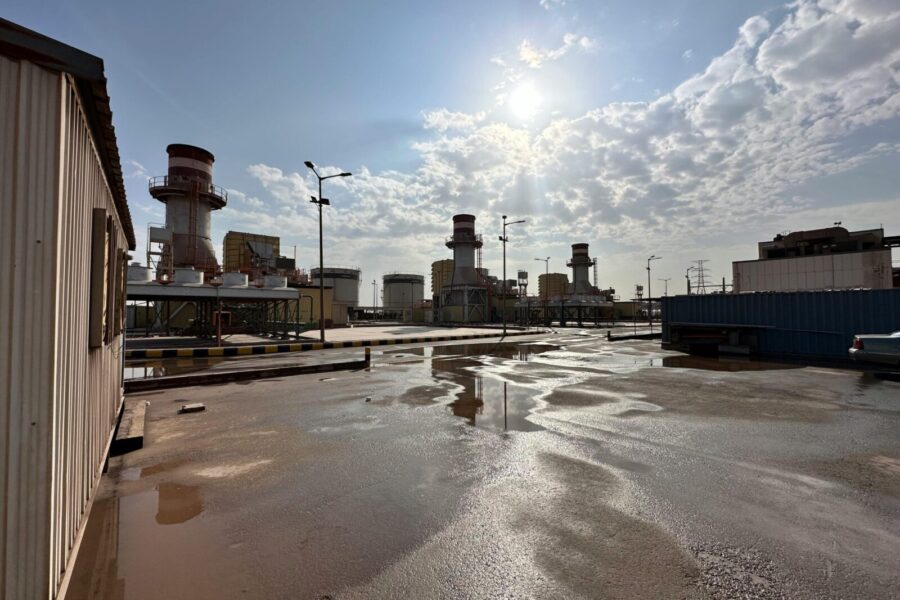 View of an industrial power plant under a cloudy sky with sunlight reflections on wet ground.