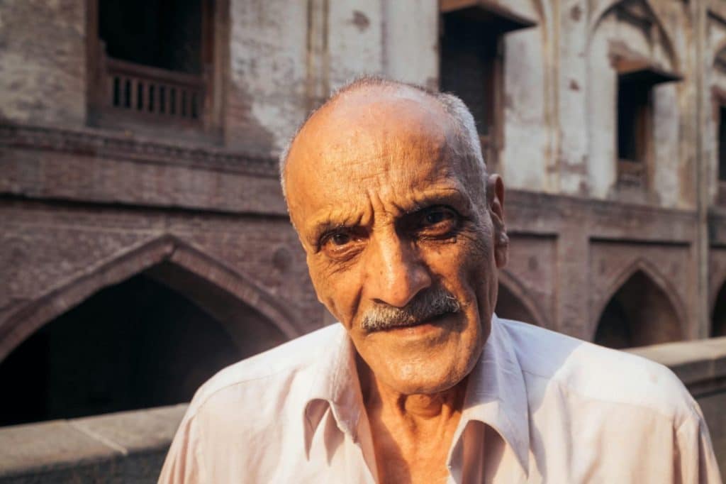 Elderly man with a mustache smiling in front of historic building in Lahore, Pakistan.