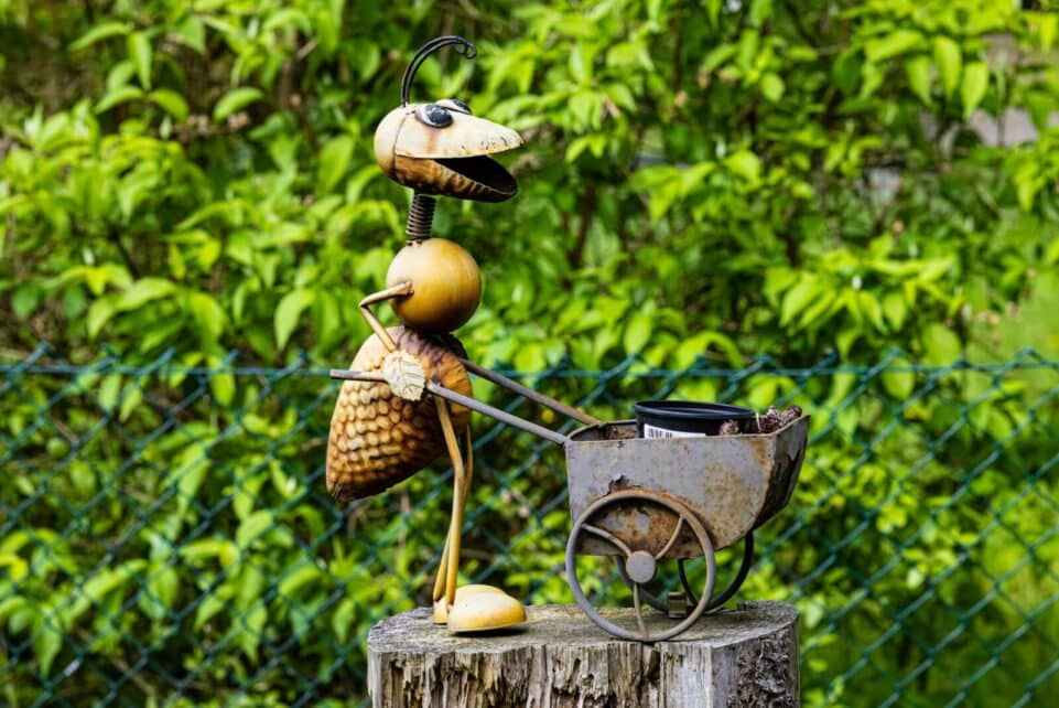 Charming metal ant sculpture with wheelbarrow in lush summer garden setting.