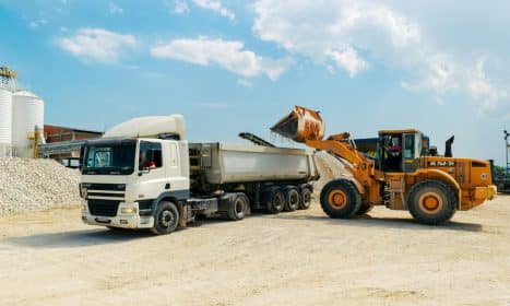 Excavator loading materials into a heavy-duty truck at a sunny construction site.