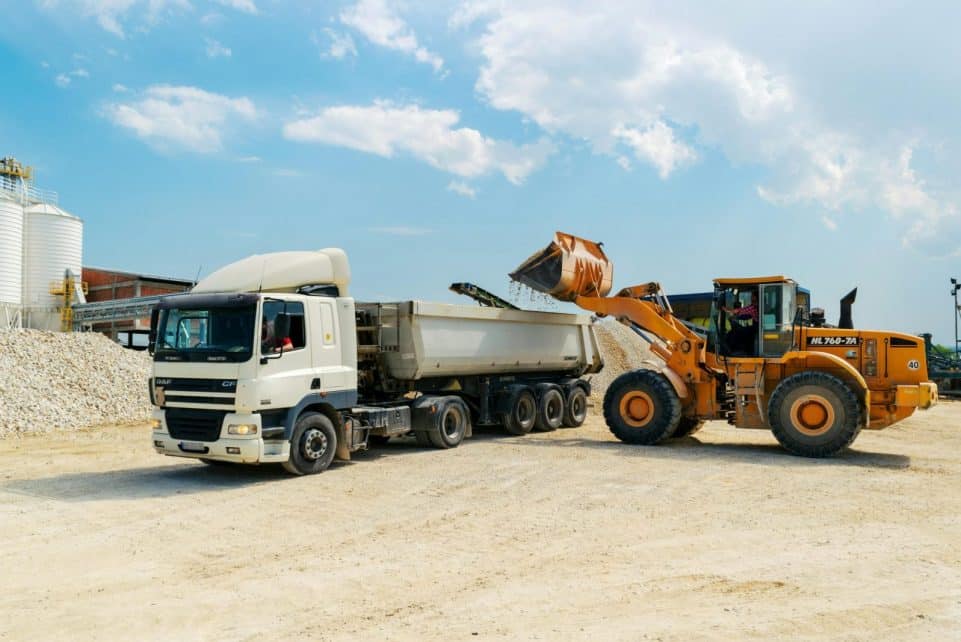 Excavator loading materials into a heavy-duty truck at a sunny construction site.