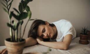 A woman peacefully resting her head on a table surrounded by indoor plants.