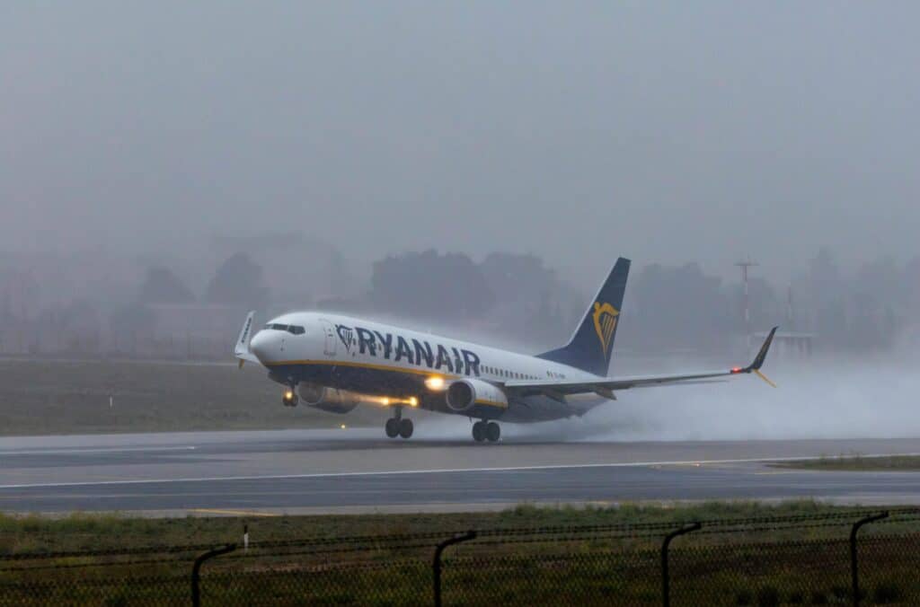 Ryanair aircraft landing on a wet runway in foggy weather creating dramatic water spray effect.