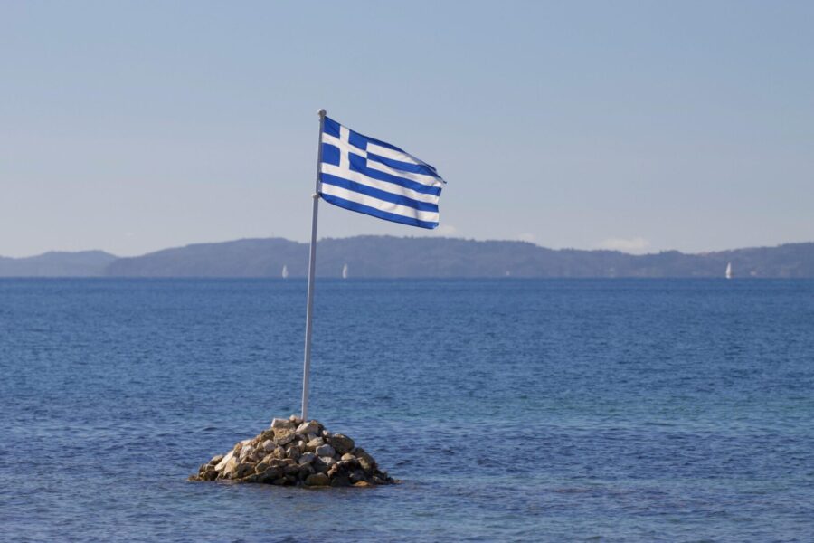 Greek flag waves over rocky island with serene Ionian Sea backdrop in Corfu, Greece.