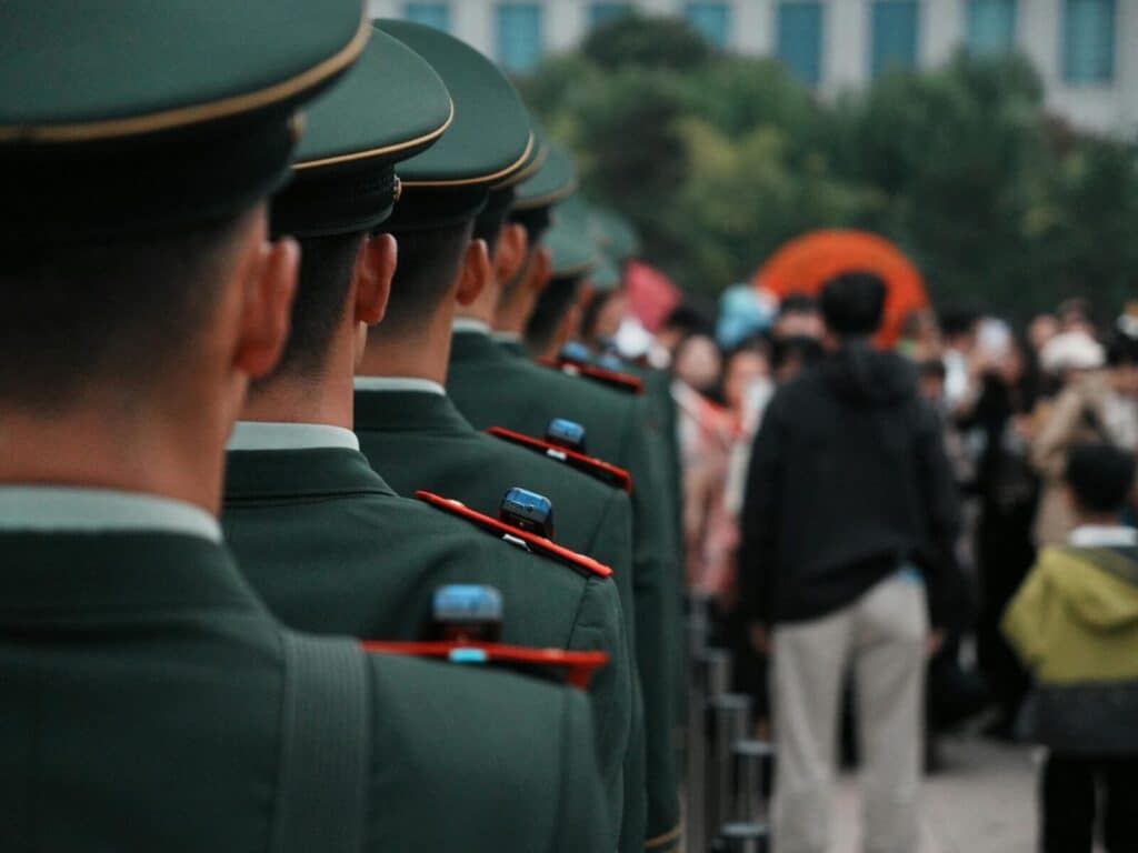 Military officers in green uniforms stand in line formation outdoors, facing a crowd.