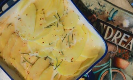 Close-up of thinly sliced potatoes with herbs and seasoning in a blue enamel dish, ready for baking.