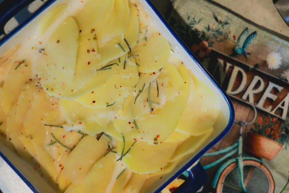 Close-up of thinly sliced potatoes with herbs and seasoning in a blue enamel dish, ready for baking.