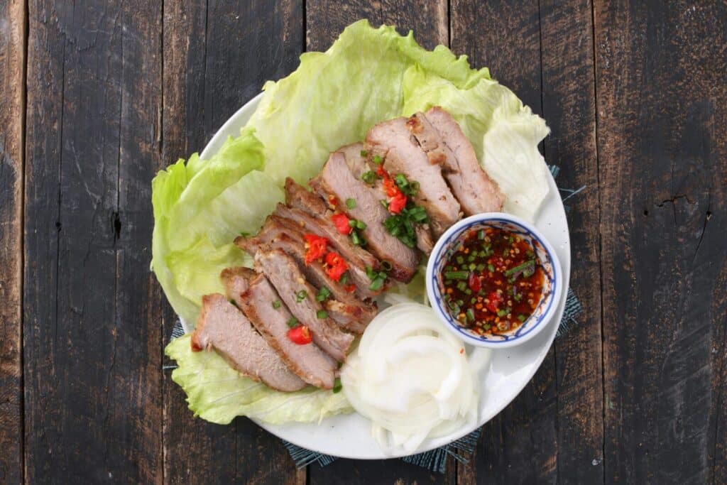 Plate of sliced grilled pork on lettuce with onions and spicy dipping sauce, presented on rustic wooden table.