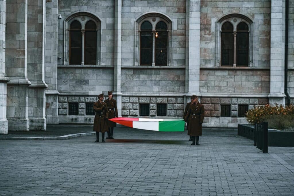 Three Hungarian soldiers stand solemnly holding a national flag in front of a historic building in Budapest.