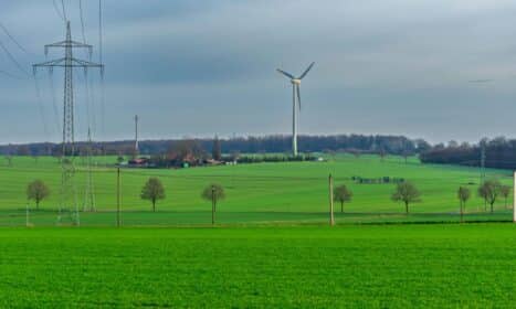 A tranquil landscape featuring green fields, a wind turbine, and power lines under an overcast sky.