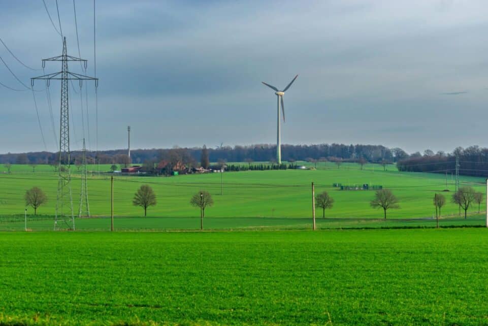 A tranquil landscape featuring green fields, a wind turbine, and power lines under an overcast sky.