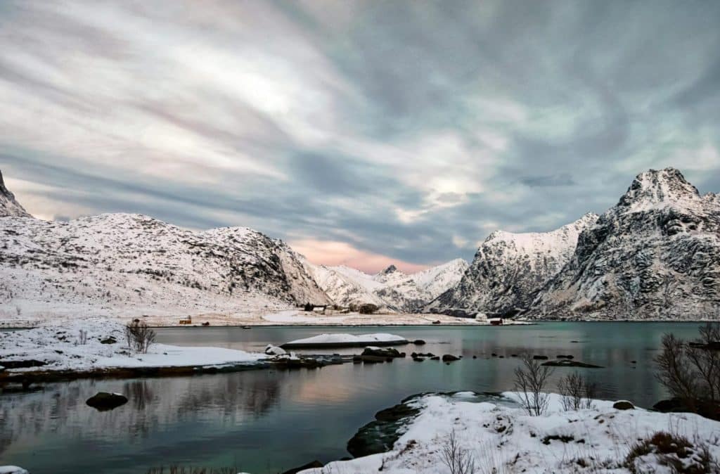 Scenic winter view of Flakstad, Norway with snow-covered mountains, fjord, and dramatic sky.
