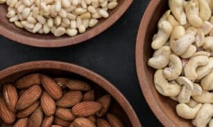 Top view of almonds, cashews, and pine nuts in wooden bowls against a dark backdrop.