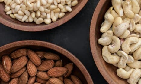 Top view of almonds, cashews, and pine nuts in wooden bowls against a dark backdrop.