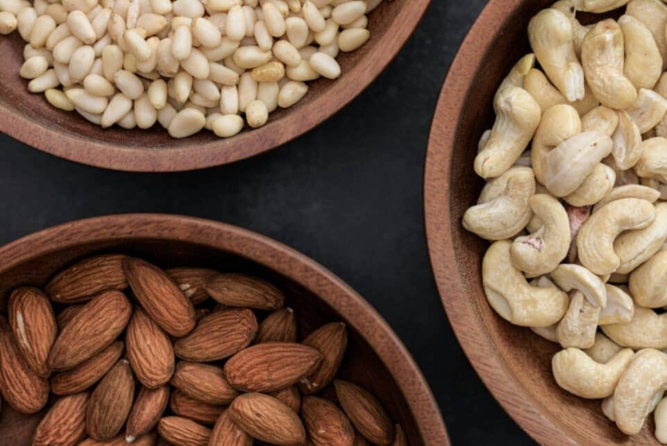 Top view of almonds, cashews, and pine nuts in wooden bowls against a dark backdrop.