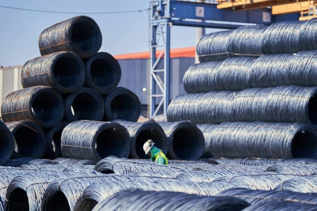 A solitary worker in protective gear inspects stacked steel coils at an industrial site.