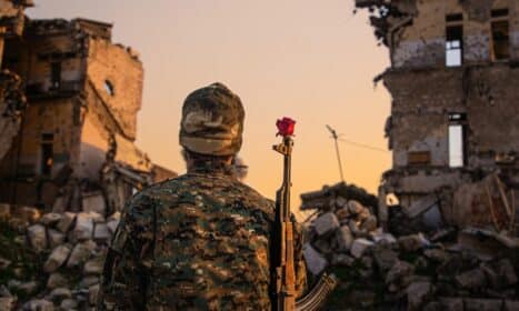 A soldier in camouflage with a red rose on rifle amidst ruined buildings at sunset.