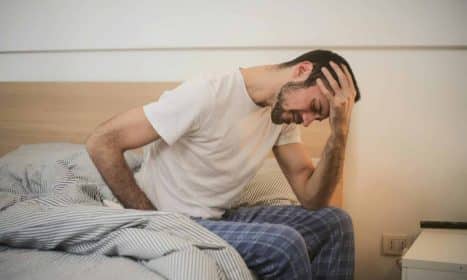 A young man in pajamas holding his head, sitting on a bed, appears to be experiencing a headache.