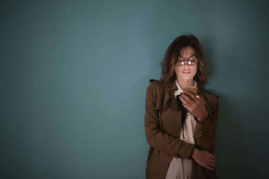 A woman with eyeglasses and brown hair uses a smartphone against a teal wall indoors.