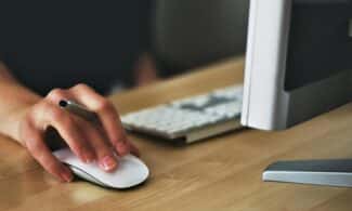 A hand using a wireless mouse at a modern desk setup with a computer and keyboard.