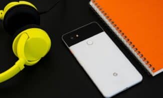 A vibrant flat lay image of a smartphone, yellow headphones, and an orange spiral notebook on a desk.