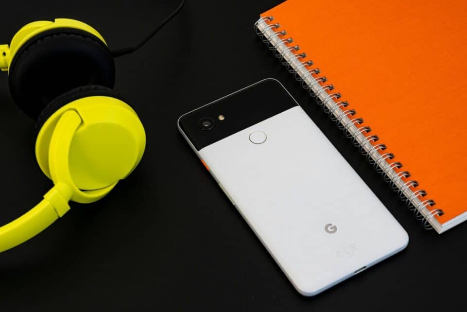 A vibrant flat lay image of a smartphone, yellow headphones, and an orange spiral notebook on a desk.