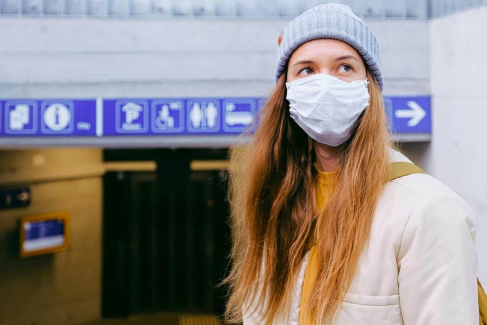 Woman in a face mask and beanie standing outdoors with signs overhead.