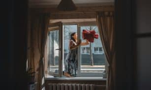 A woman cleaning a window in a well-designed cozy apartment with curtains and natural light.