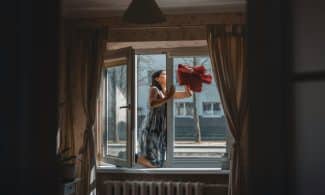 A woman cleaning a window in a well-designed cozy apartment with curtains and natural light.