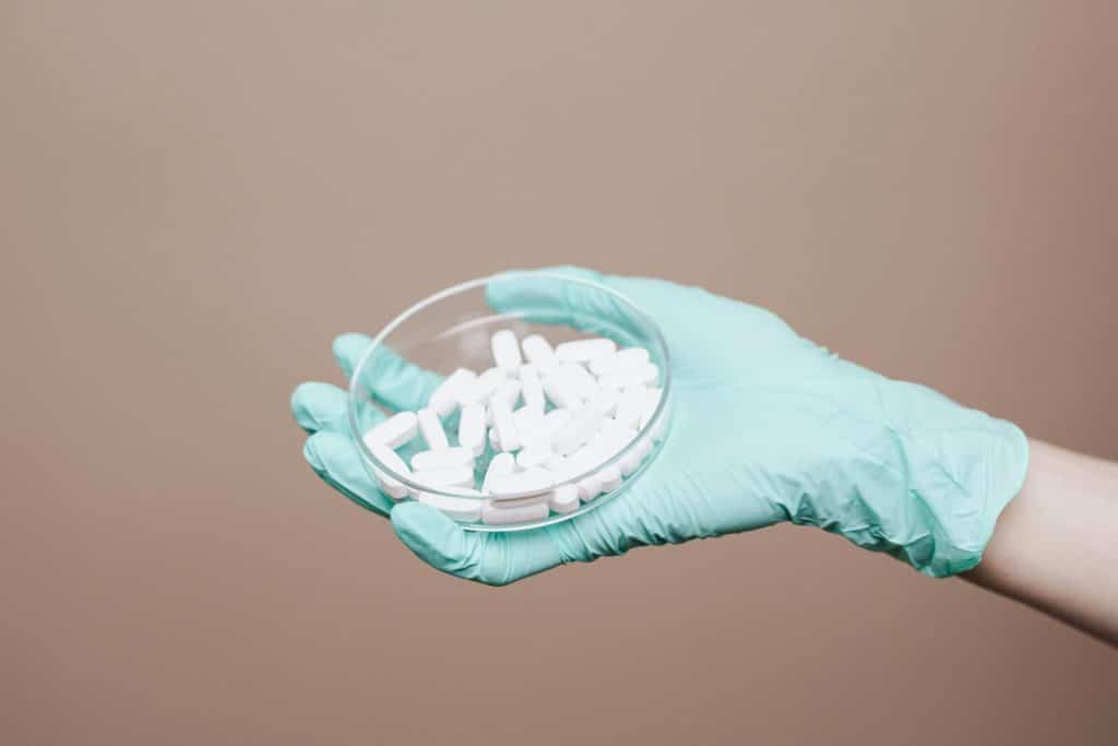 A gloved hand holding a petri dish filled with white pills in a healthcare setting.