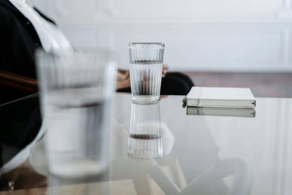 A minimalist composition featuring a glass of water and notebook on a reflective glass table.