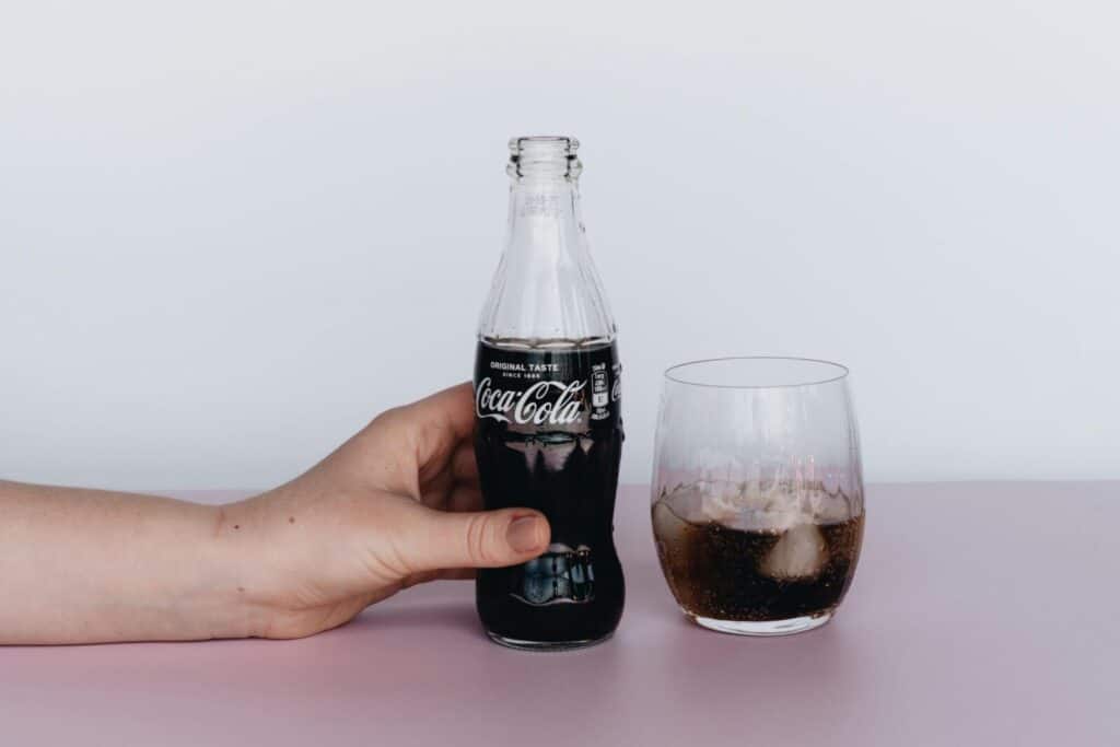 Close-up of a hand holding a Coca-Cola bottle next to a glass filled with ice and soda.