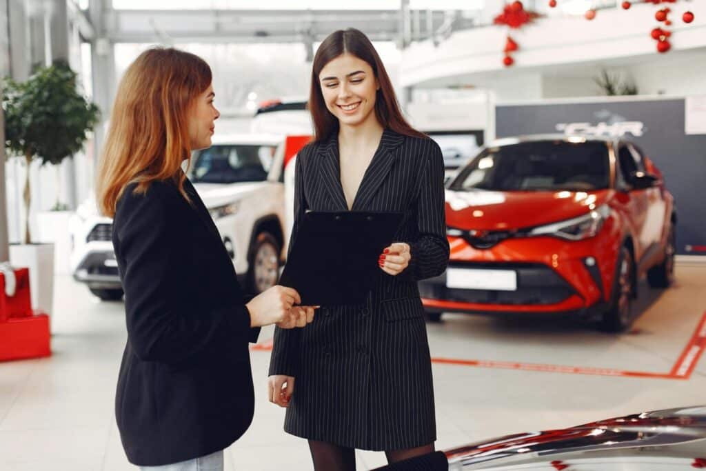Cheerful young friendly dealer in formal stylish black dress showing contract to smiling female customer in black jacket while standing in car showroom against new shiny automobiles