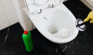 From above of crop anonymous person in rubber gloves using liquid toilet sanitizer and brush to clean toilet bowl in bathroom